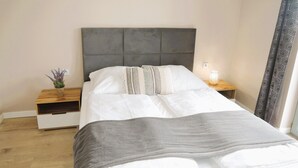 Una cama bien hecha con cabecera gris, una mesita de noche con lámpara y una planta pequeña en el buró.