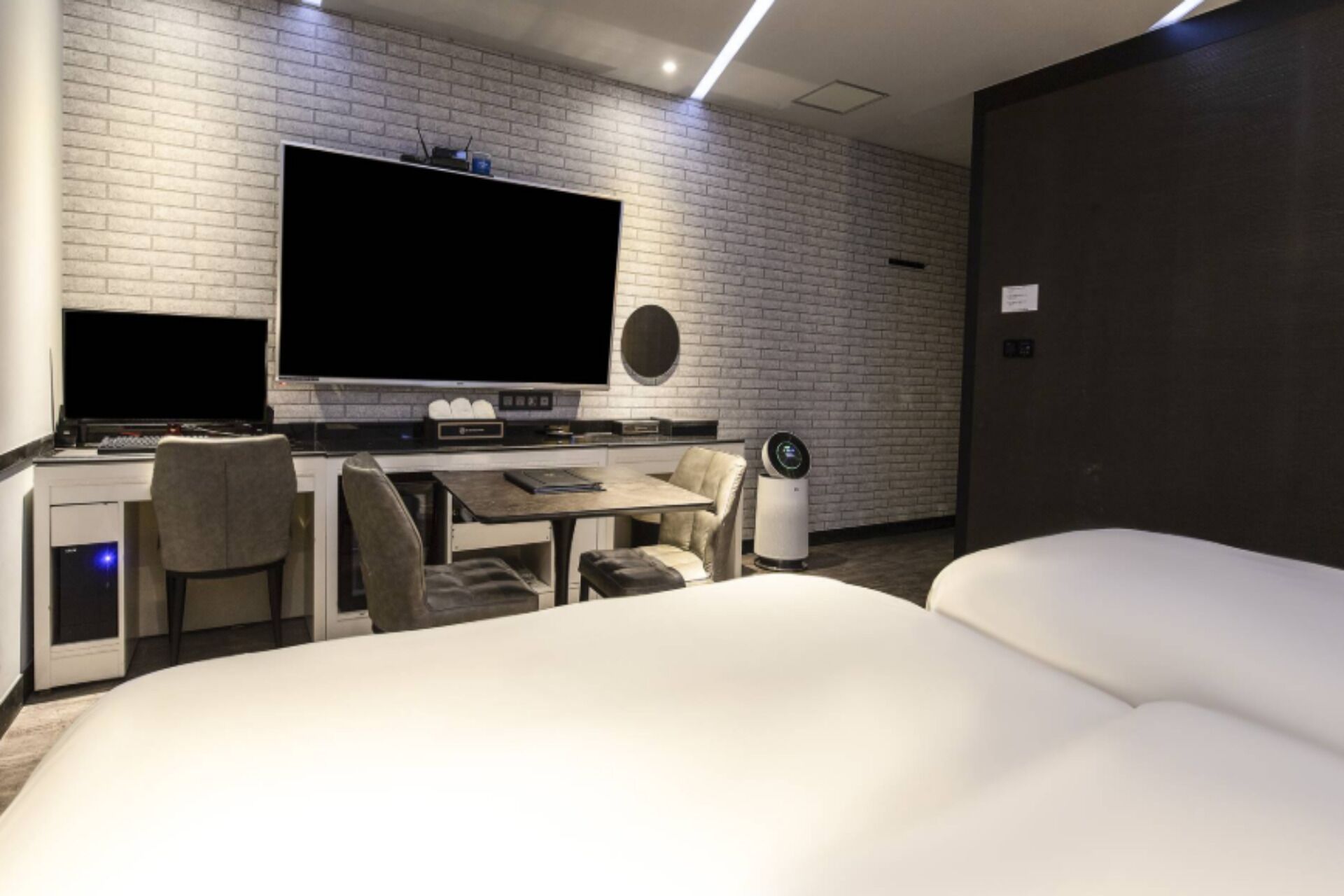 Una habitación de hotel moderna con un televisor de pantalla plana grande montado en una pared de ladrillo, un escritorio con una computadora, dos sillas y una cama con ropa blanca.