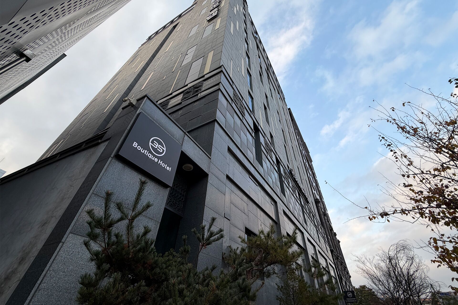 A tall building with a Boutique Hotel sign, featuring a modern architectural design with a prominent entrance and surrounding trees.