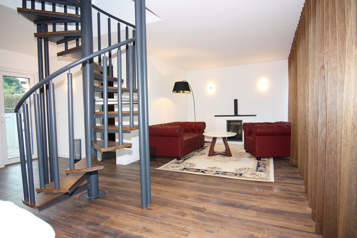 A modern living room with a spiral staircase, red sofas, a round table, and a wooden floor.