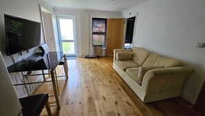 A living room with a beige sofa, a TV mounted on the wall, and a wooden floor. - Standard Single Room - Shared (Windsor)