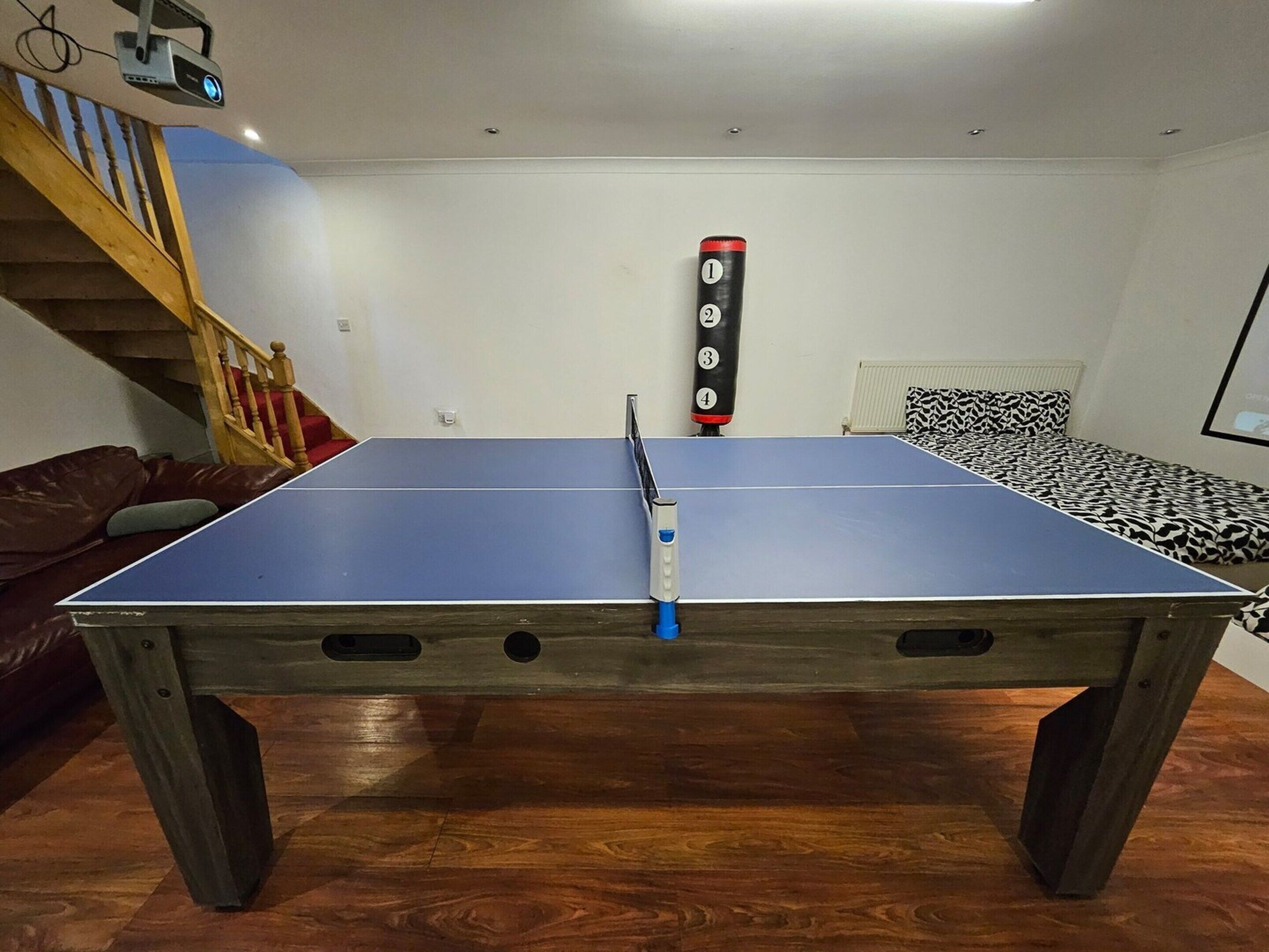 A ping pong table in a room with a wooden floor, a staircase, and a bed with patterned bedding.