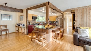 A spacious living area with a wooden floor, a central island with stools, a sofa, and a kitchen in the background. - Lodge at Deception Pass | Trails, Hot Tub, Walk to Cornet Bay (Oak Harbor)