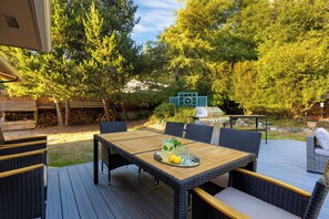An outdoor patio with a wooden table and wicker chairs, surrounded by trees and a barbecue grill. - Lodge at Deception Pass | Trails, Hot Tub, Walk to Cornet Bay (Oak Harbor)
