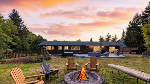 A backyard with a fire pit, wooden chairs, and a modern house in the background. - Lodge at Deception Pass | Trails, Hot Tub, Walk to Cornet Bay (Oak Harbor)