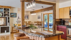 A kitchen with a large island, wooden cabinets, and a dining area with chairs. - Lodge at Deception Pass | Trails, Hot Tub, Walk to Cornet Bay (Oak Harbor)