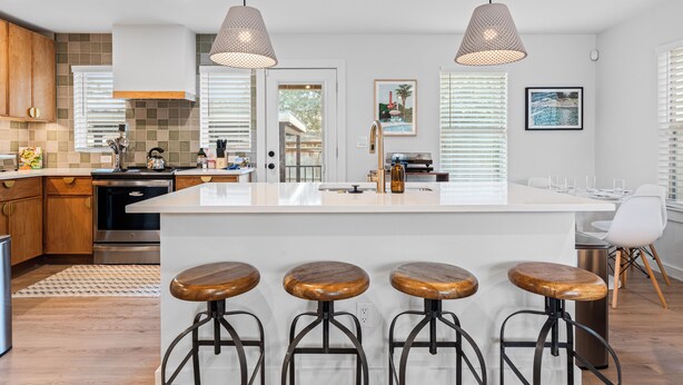 A modern kitchen with a central island, wooden bar stools, pendant lighting, and a dining area with a white chair. - Casa Que Canta by AvantStay | Private Pool, Firepit & Walkable East Austin Gem (Austin)