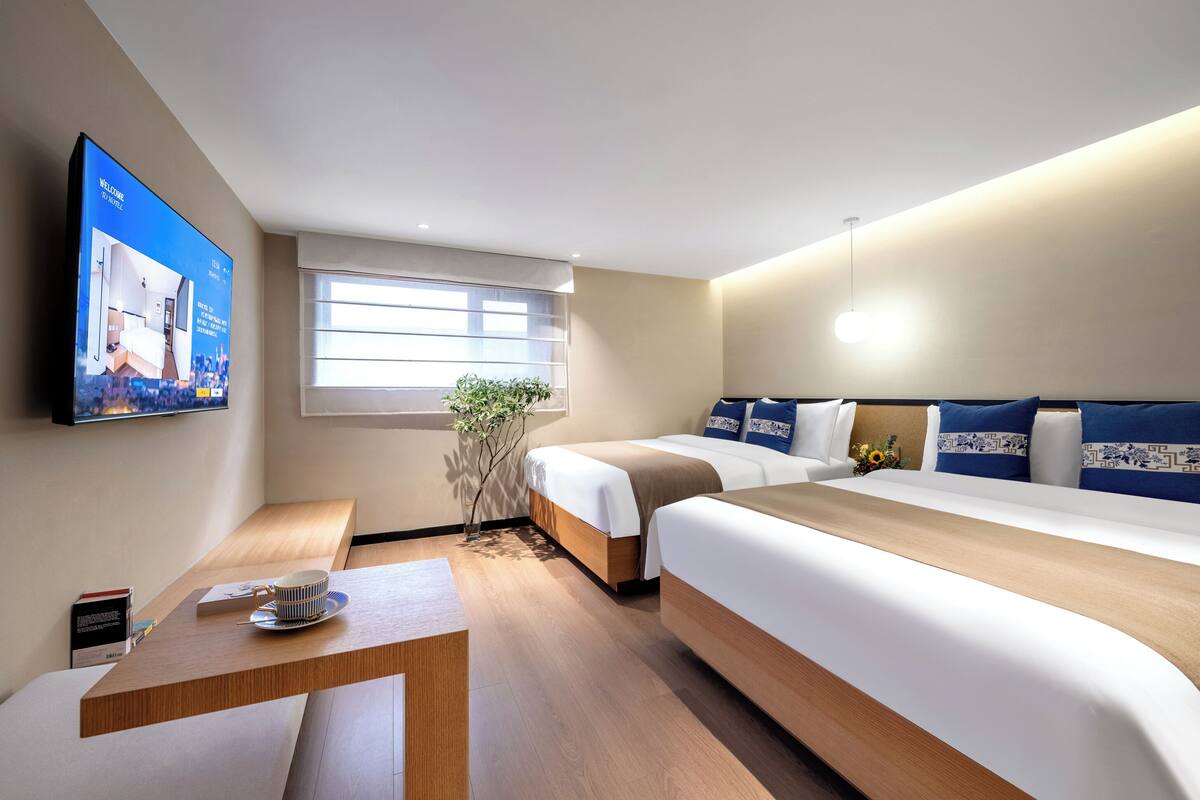 Habitación de hotel con dos camas, un televisor en la pared, una ventana y una mesita con una taza y un plato.