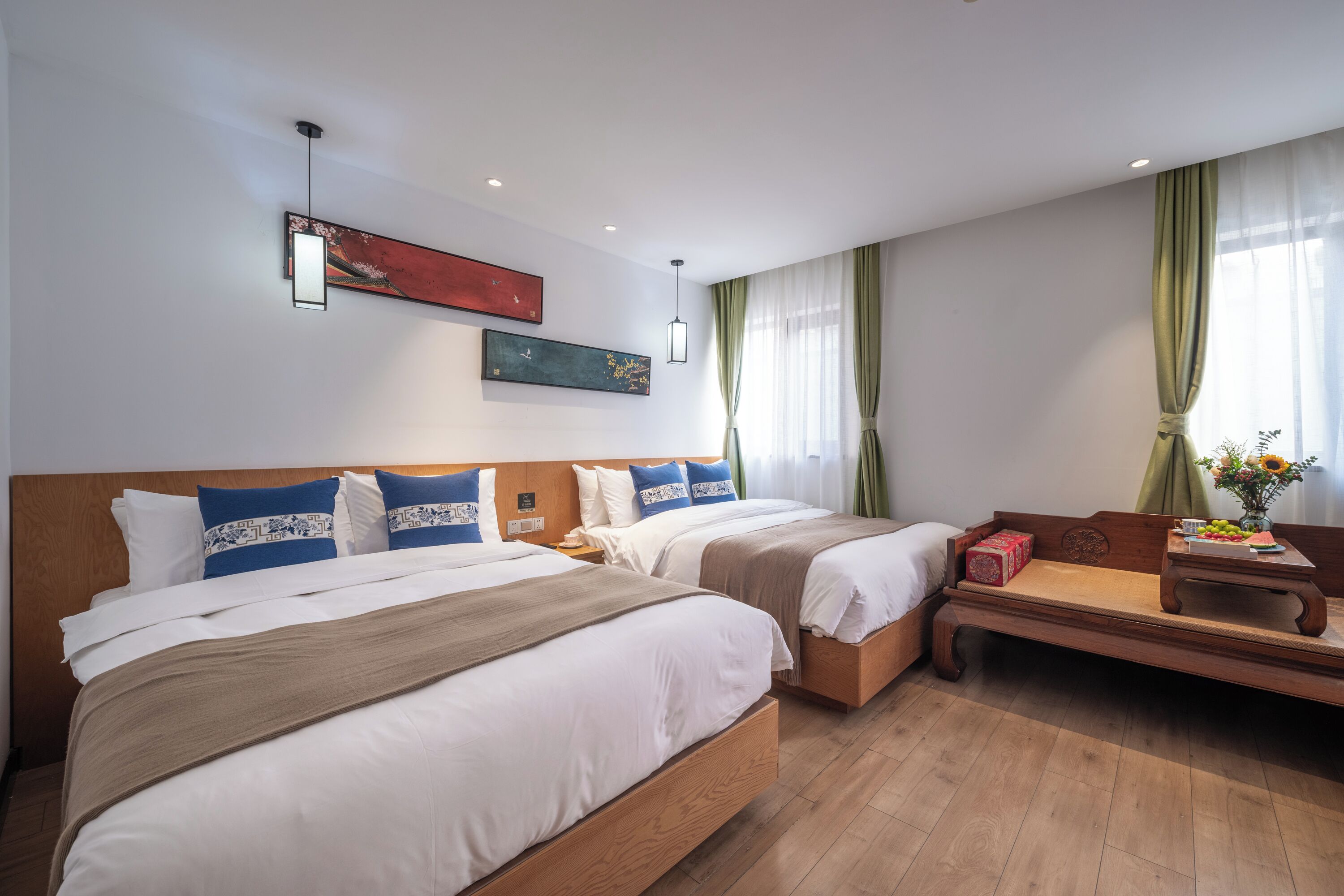 Una habitación de hotel con dos camas, un banco de madera, una mesita con un florero y obras de arte en las paredes.