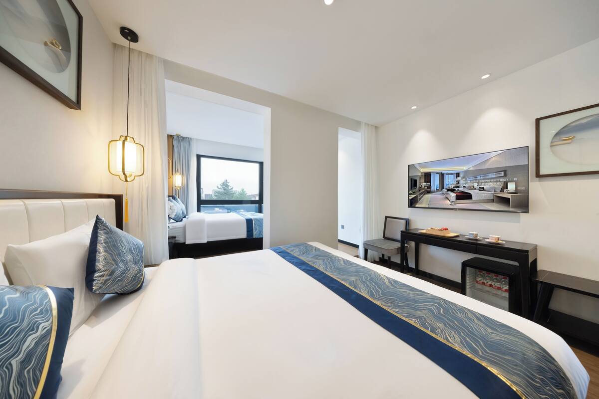 Habitación de hotel moderna con una cama grande, un escritorio, una silla, un refrigerador pequeño y un ventanal con vistas al exterior.