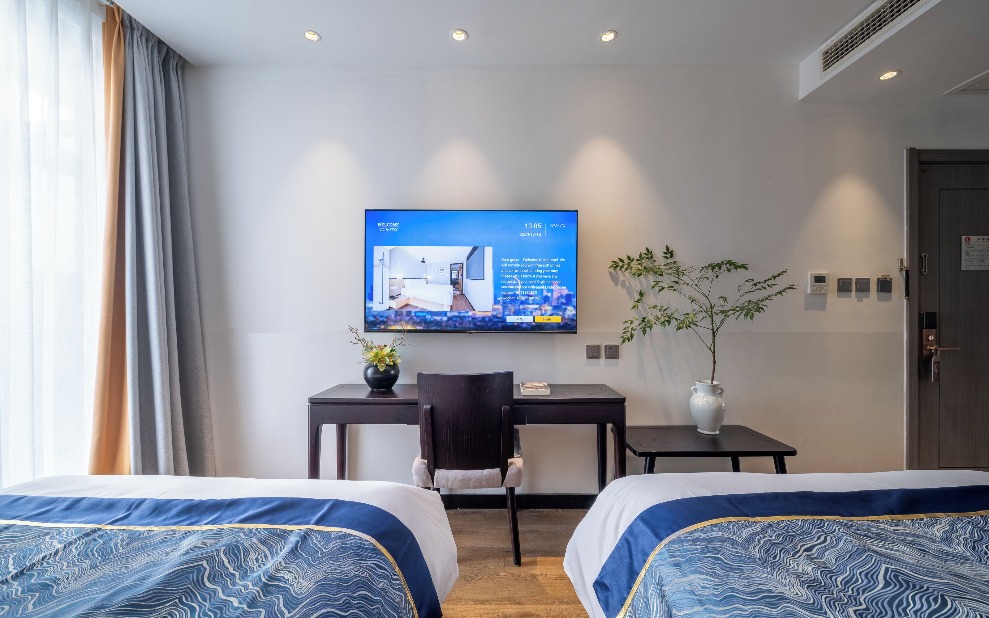 A hotel room with two beds, a desk, a chair, a television displaying a welcome message, a plant, and a door with a sign.