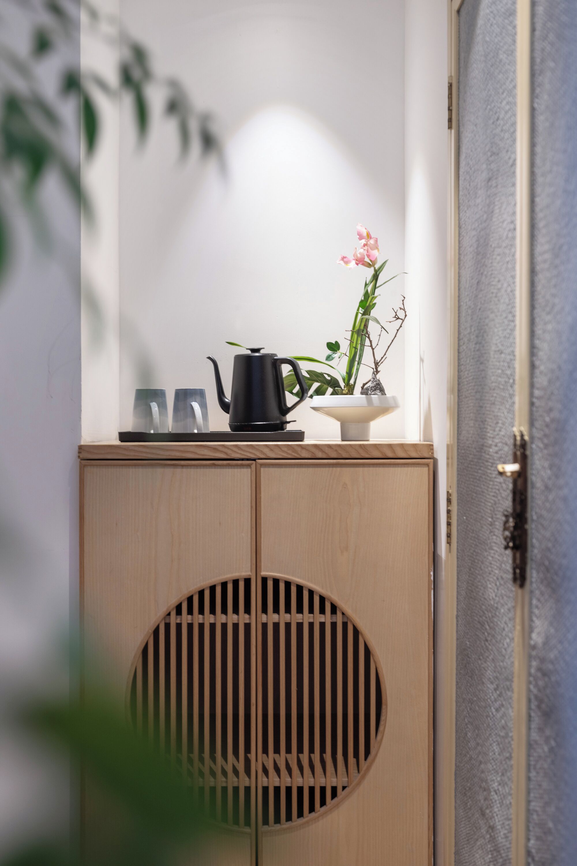 A wooden cabinet with a circular cutout, a black teapot, two glasses, and a potted plant with pink flowers.