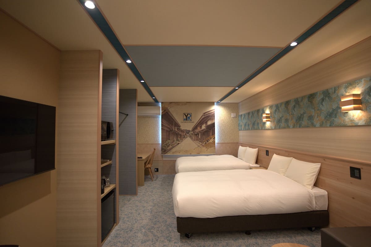 A hotel room with two beds, a desk, a chair, a TV, and a mural on the wall.