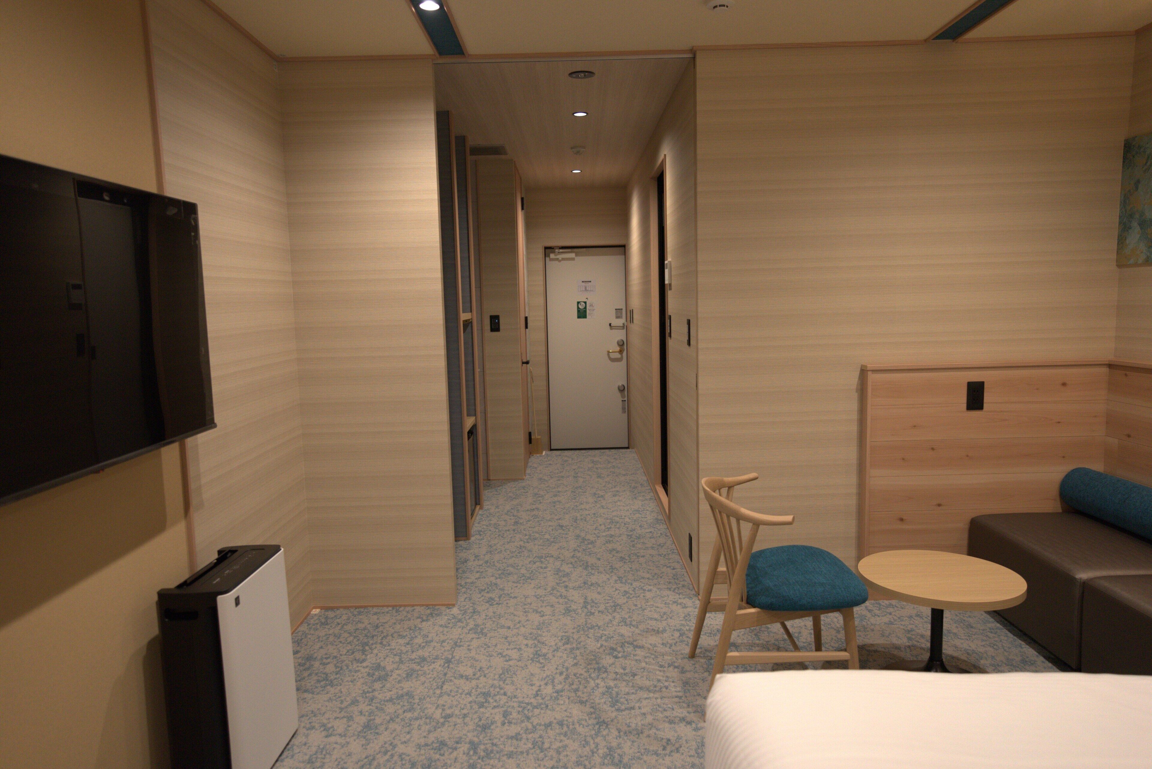 Habitación de hotel con cama, una mesita, una silla, televisor y una puerta que da a otra habitación.