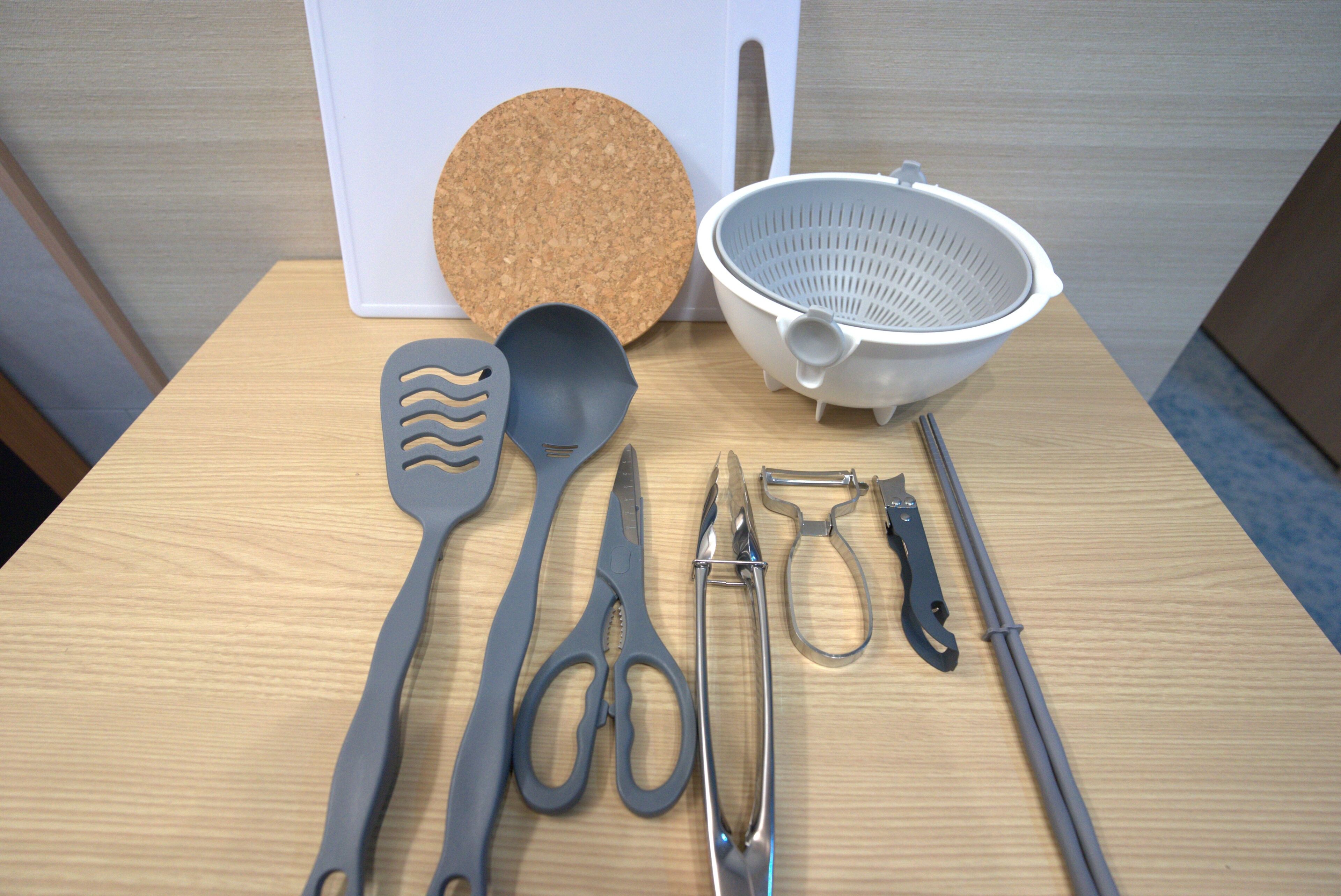 A table with kitchen utensils including a cork mat, colander, spatula, scissors, and tongs.