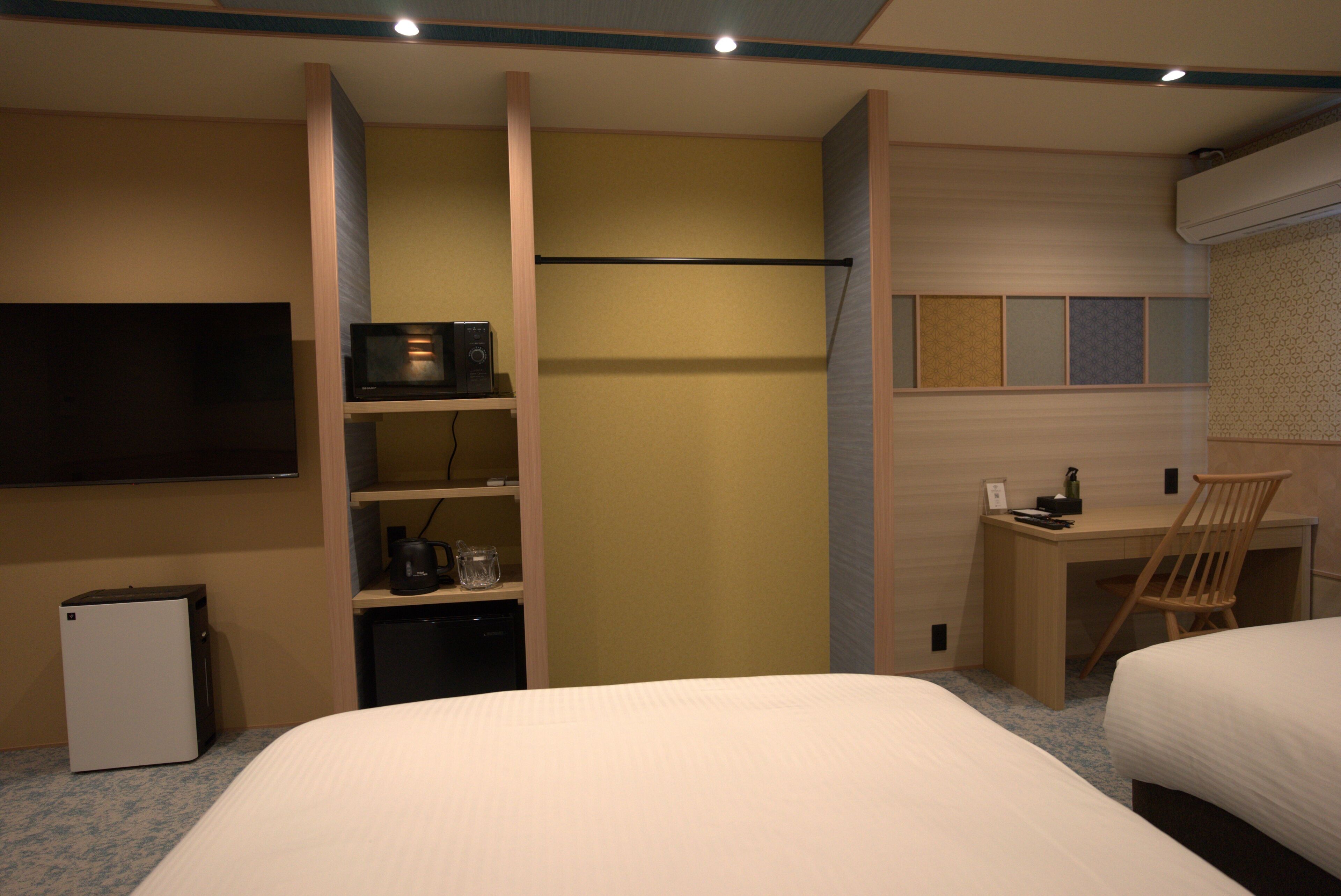 A hotel room with a bed, a desk, a chair, a TV, a mini-fridge, and a shelf with a kettle.