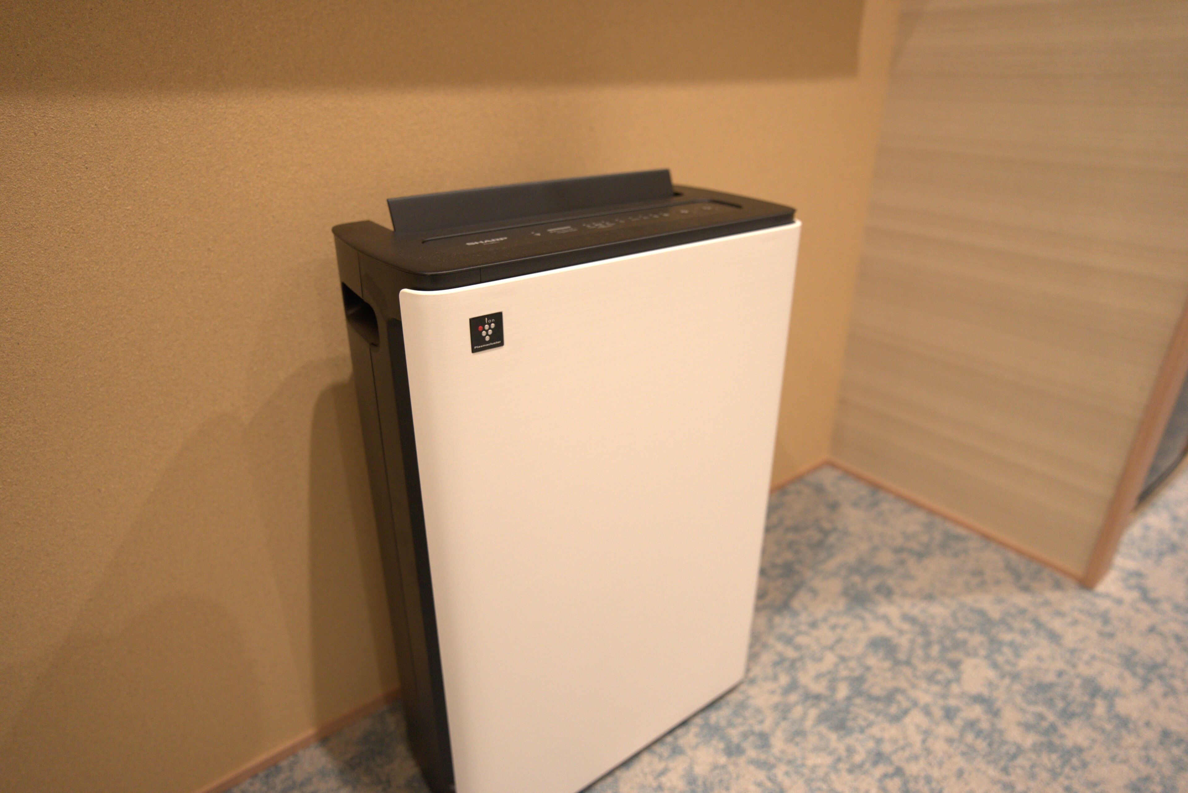 Un dispositivo blanco y compacto, fijado a la pared y con un panel de control, probablemente un termostato u otro aparato similar, en una habitación con pared lisa y suelo estampado.