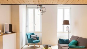 Een moderne zithoek met een bank, een blauwe fauteuil, een klein rond tafeltje en een televisie op een kast.