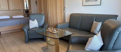 Una sala de estar moderna con una mesa de centro de vidrio, un sofá de cuero gris y una cama con cabecera de madera.
