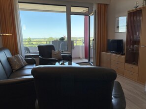 Un salon moderne avec un canapé, un fauteuil et une table basse en verre. Un téléviseur est fixé au mur, et on aperçoit un meuble sur le côté droit. La pièce est dotée d’une grande porte-fenêtre coulissante qui donne sur un balcon offrant une vue sur la verdure.