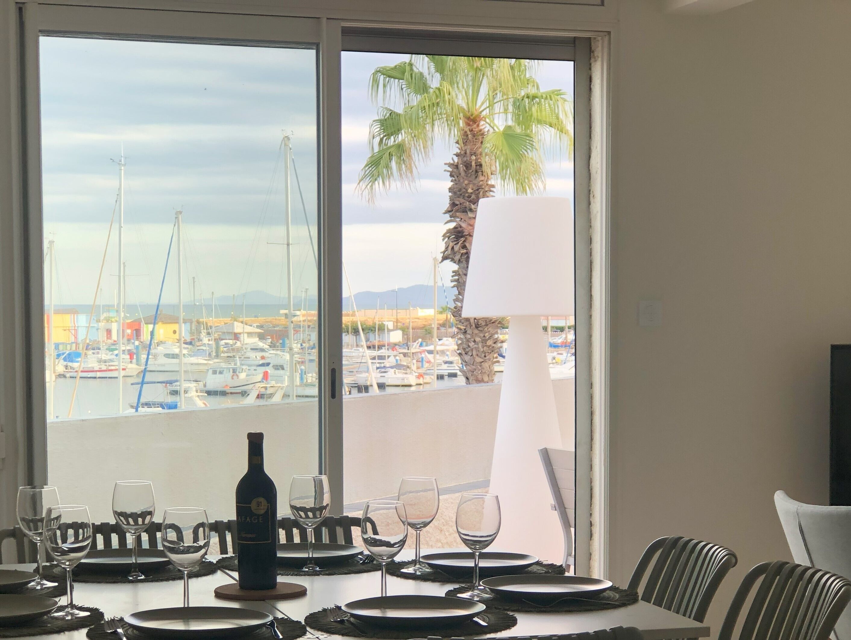 A dining table with wine glasses and a bottle, overlooking a marina with boats and a palm tree.