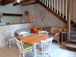 Una habitación acogedora con una mesa de madera, sillas blancas, un sofá con cojines, una escalera y un estante en la pared con objetos decorativos.