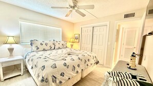 Ein Schlafzimmer mit einem Bett, einem Nachttisch, einer Lampe, einem Deckenventilator, einem Fenster mit Jalousien und einer Tür.