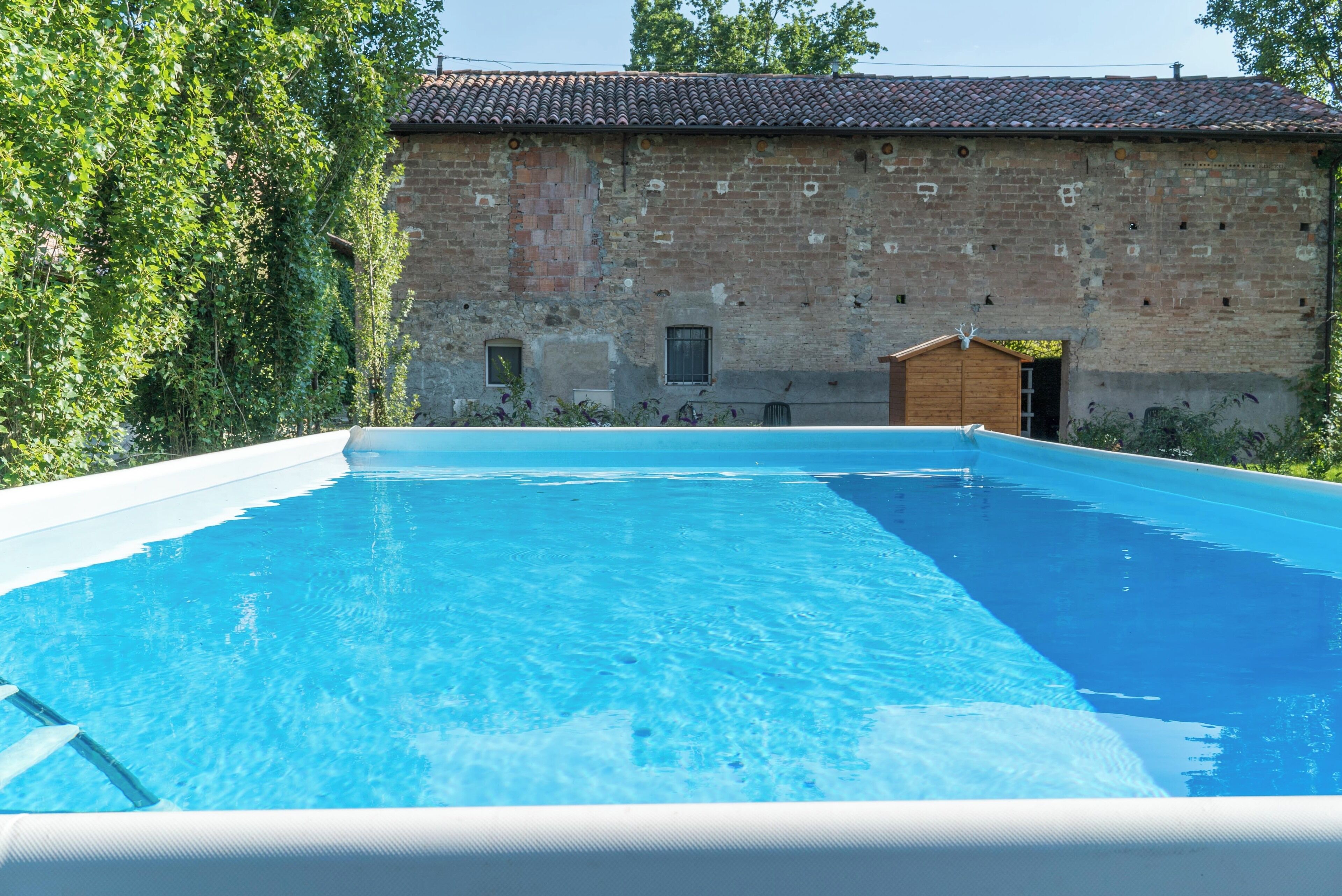 Una piscina rectangular con agua azul cristalina, rodeada por un edificio de ladrillo con techo de azulejos y una pequeña construcción de madera al fondo.