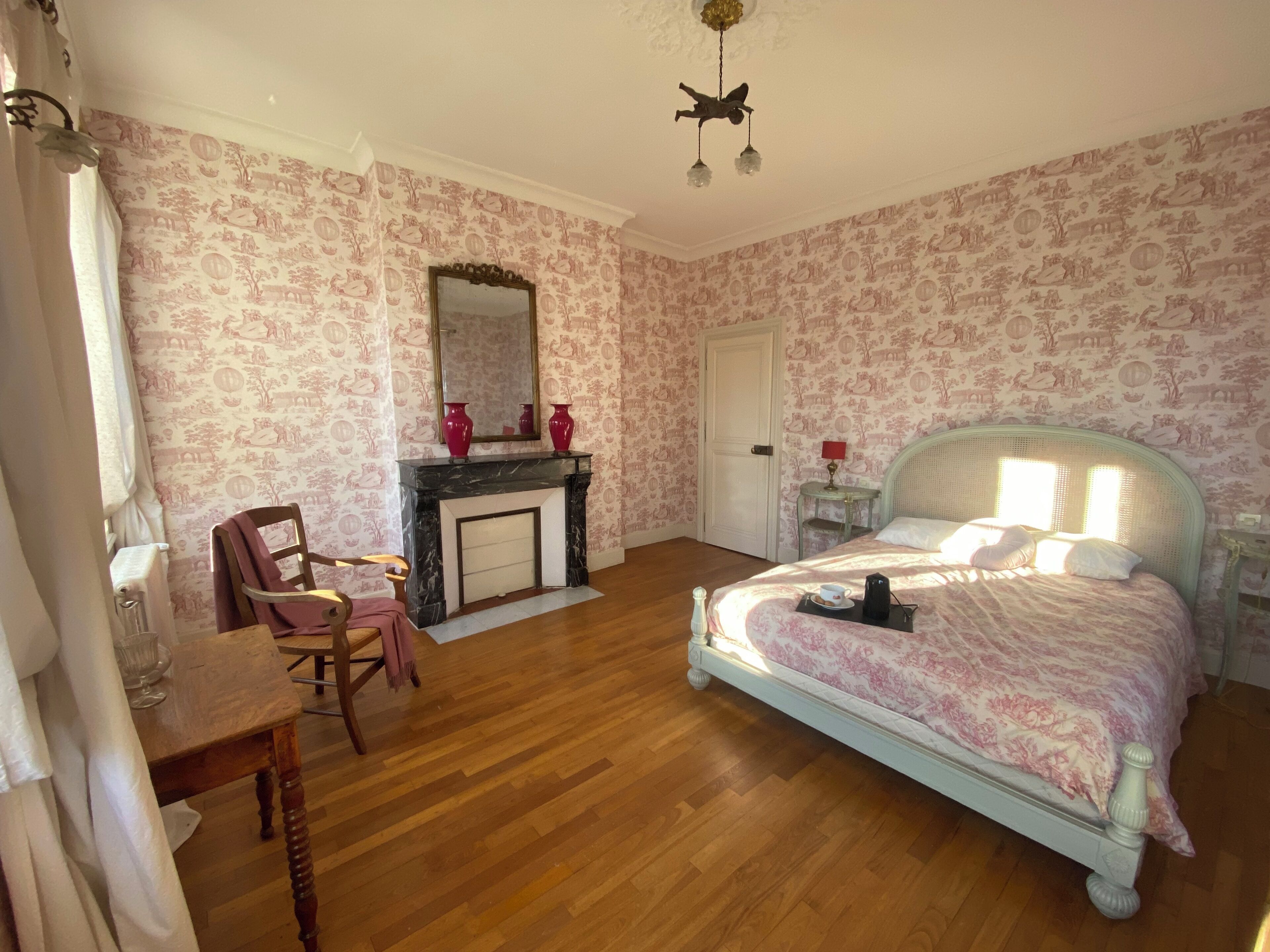 A bedroom with a bed, a chair, a table, a fireplace, and a mirror.