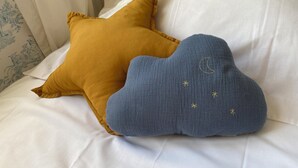A bed with white bedding and two decorative pillows, one star-shaped and one cloud-shaped, with embroidered details.