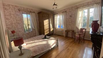 A bedroom with a bed, a chair, a table, a radiator, and a window with curtains.