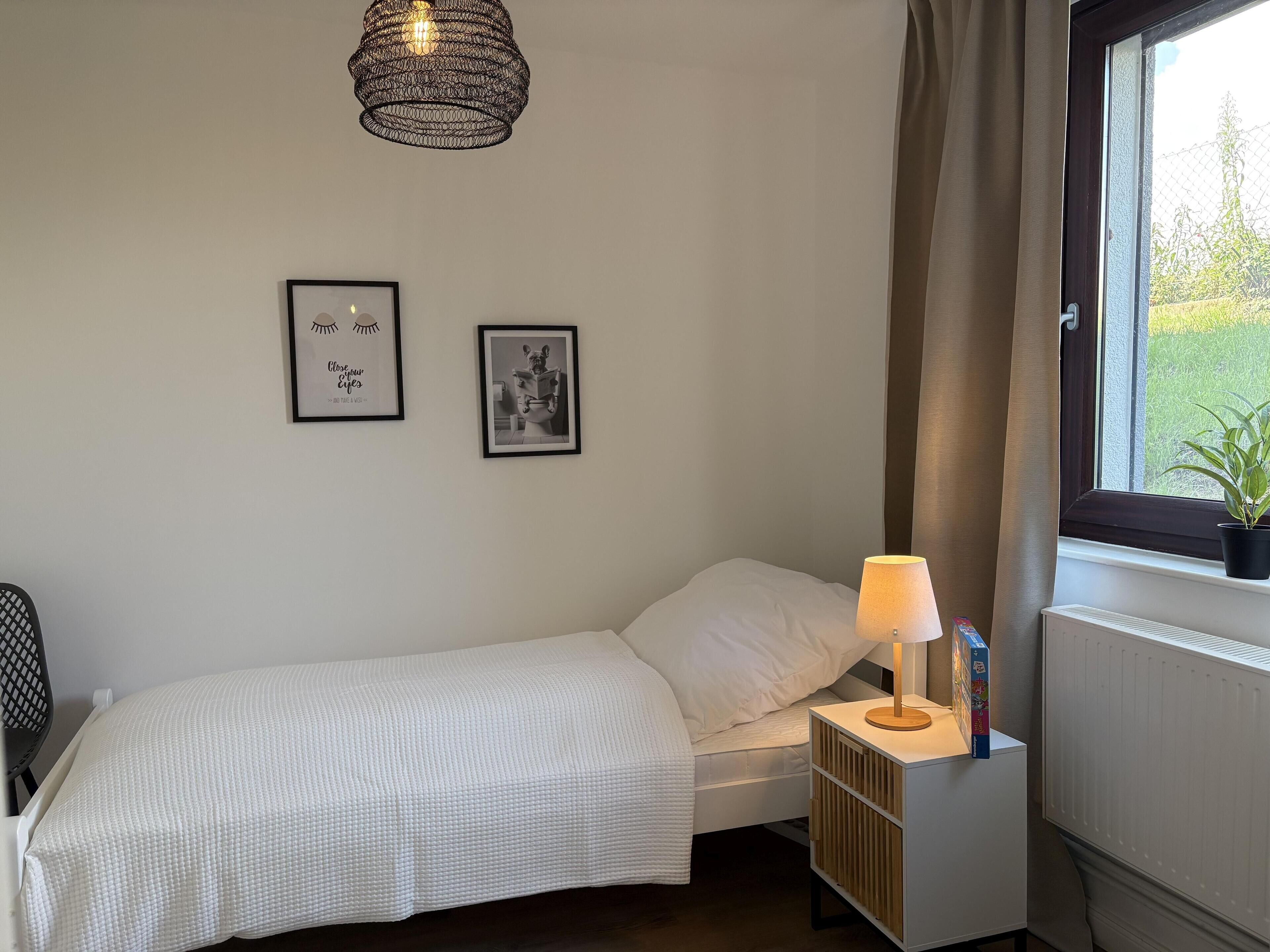 A bedroom with a single bed, a bedside table with a lamp, a radiator, and a window with a view of greenery.