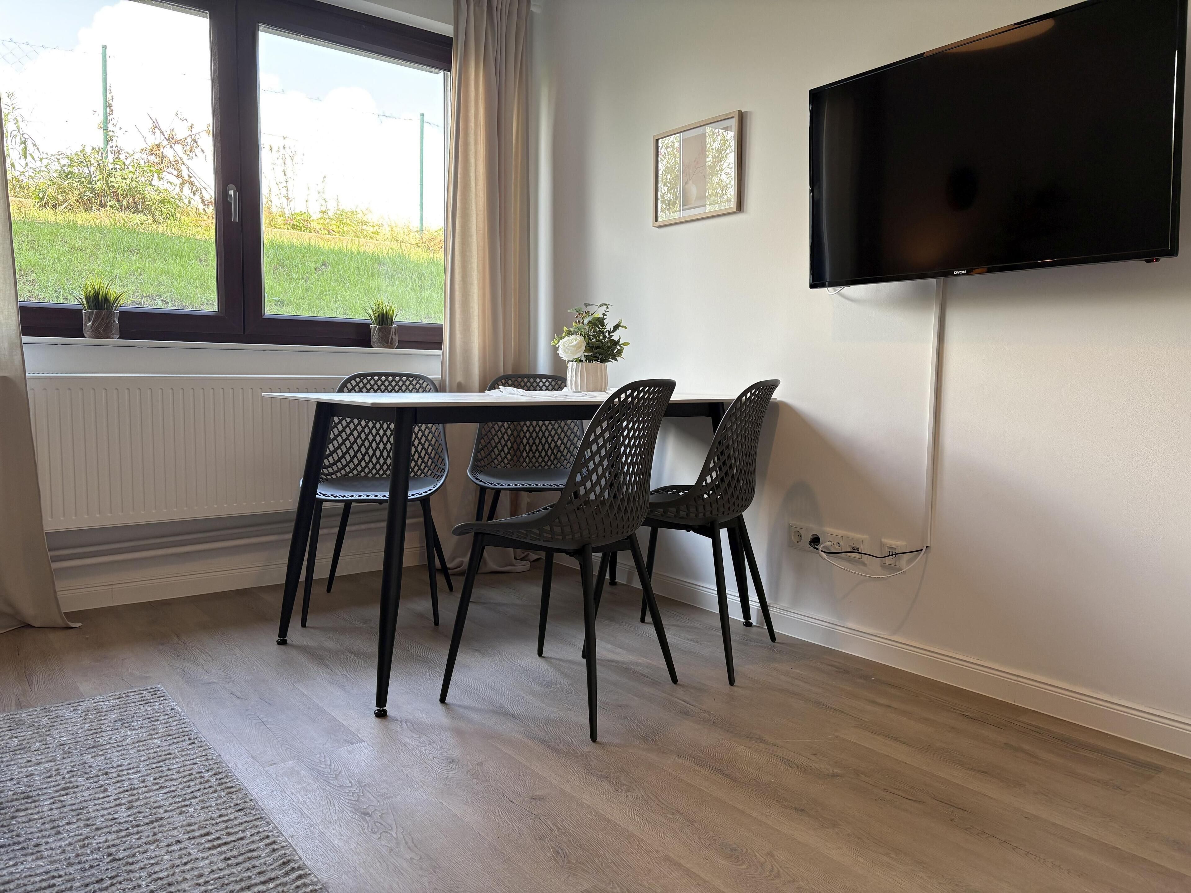 Ein Essbereich mit Tisch und Stühlen, ein Fenster mit Blick auf Grünflächen, eine Heizung und ein an der Wand befestigter Fernseher.