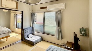 Ein traditioneller japanischer Raum mit Tatami-Böden, einer Futon-Liege, einem niedrigen Holztisch, einem Fernseher und einer Klimaanlage.