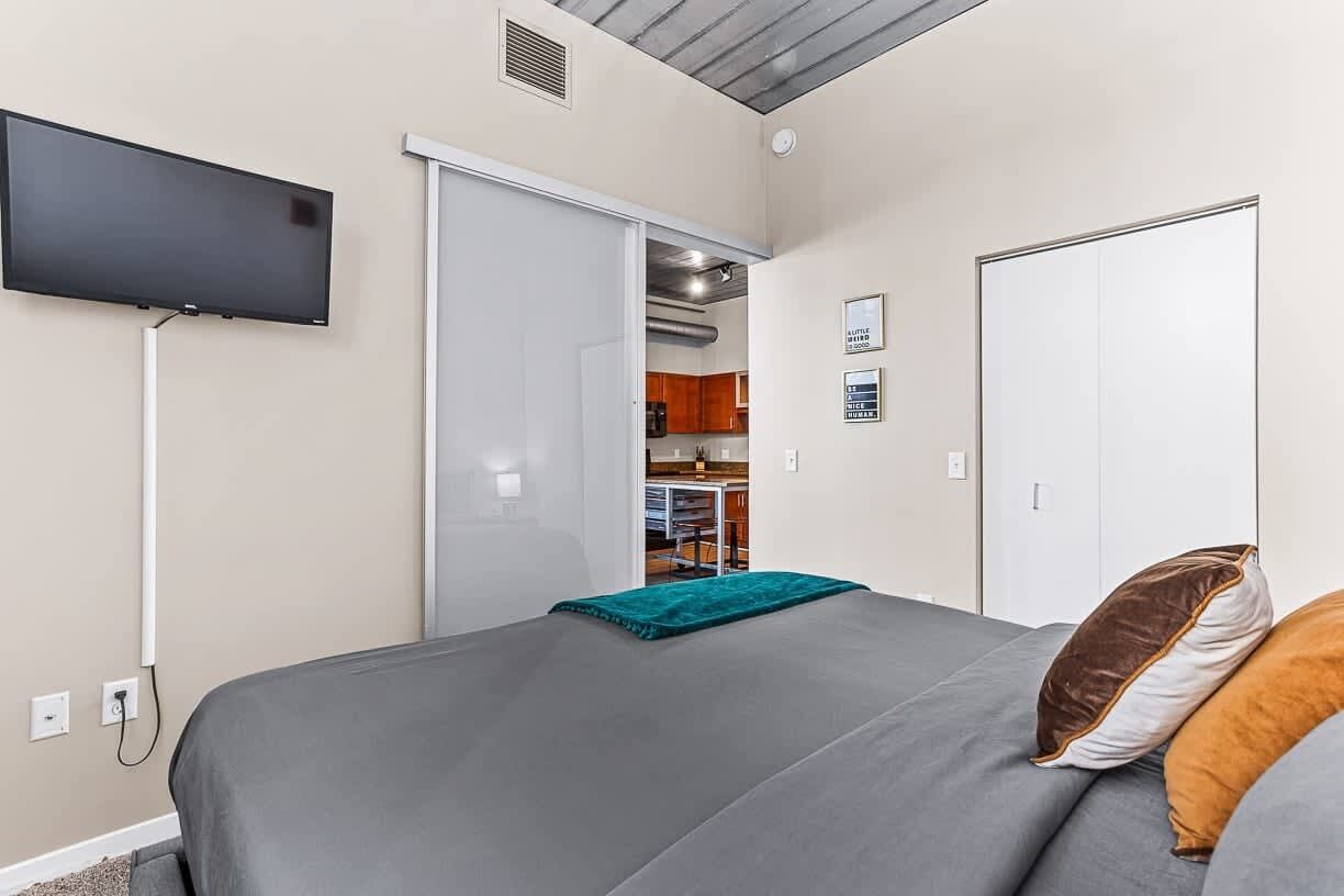 Un dormitorio con cama, televisor montado en la pared, armario y una zona de cocina visible a través de una puerta abierta.