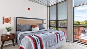 A modern bedroom with a large bed, a nightstand, a potted plant, and a view of the outside through large windows.