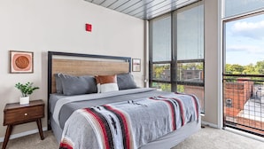 A modern bedroom with a large bed, a nightstand, a potted plant, and a view of the outside through large windows.