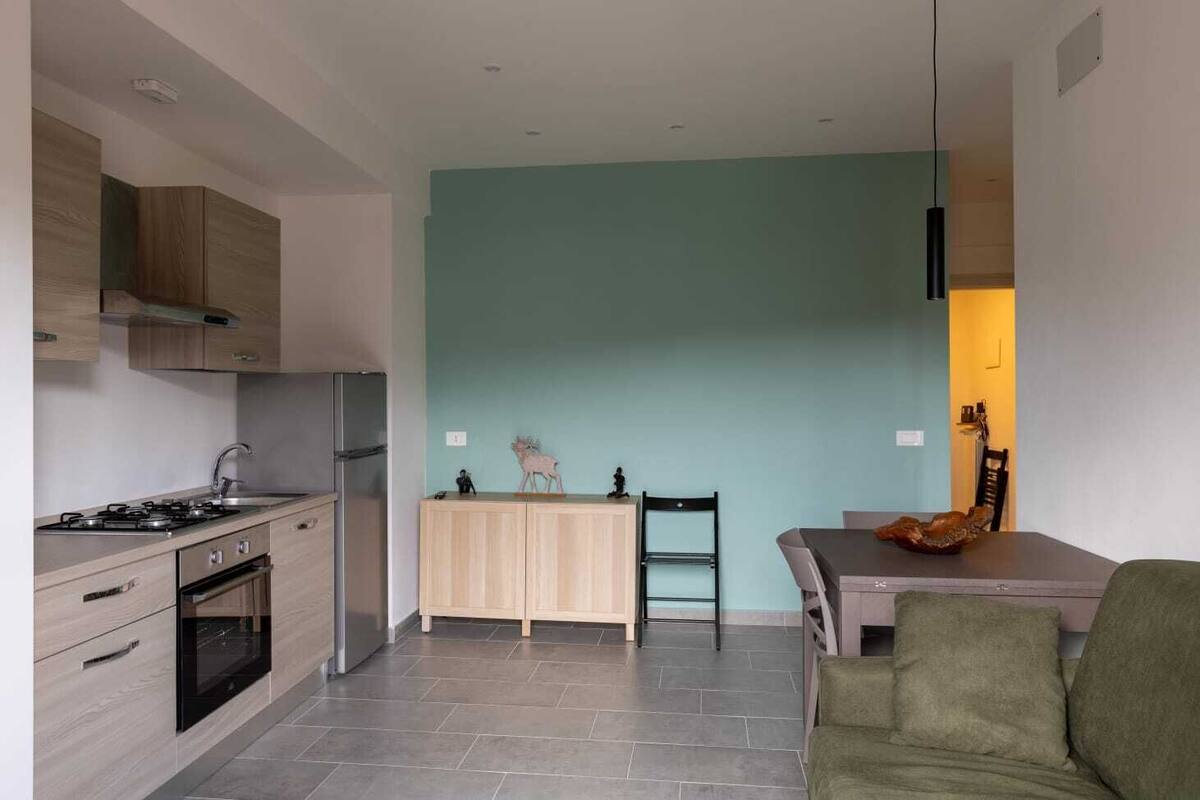 Una cucina moderna con piano cottura, forno e frigorifero integrati. C'è un'area pranzo con tavolo e sedie, e un divano verde nel soggiorno.