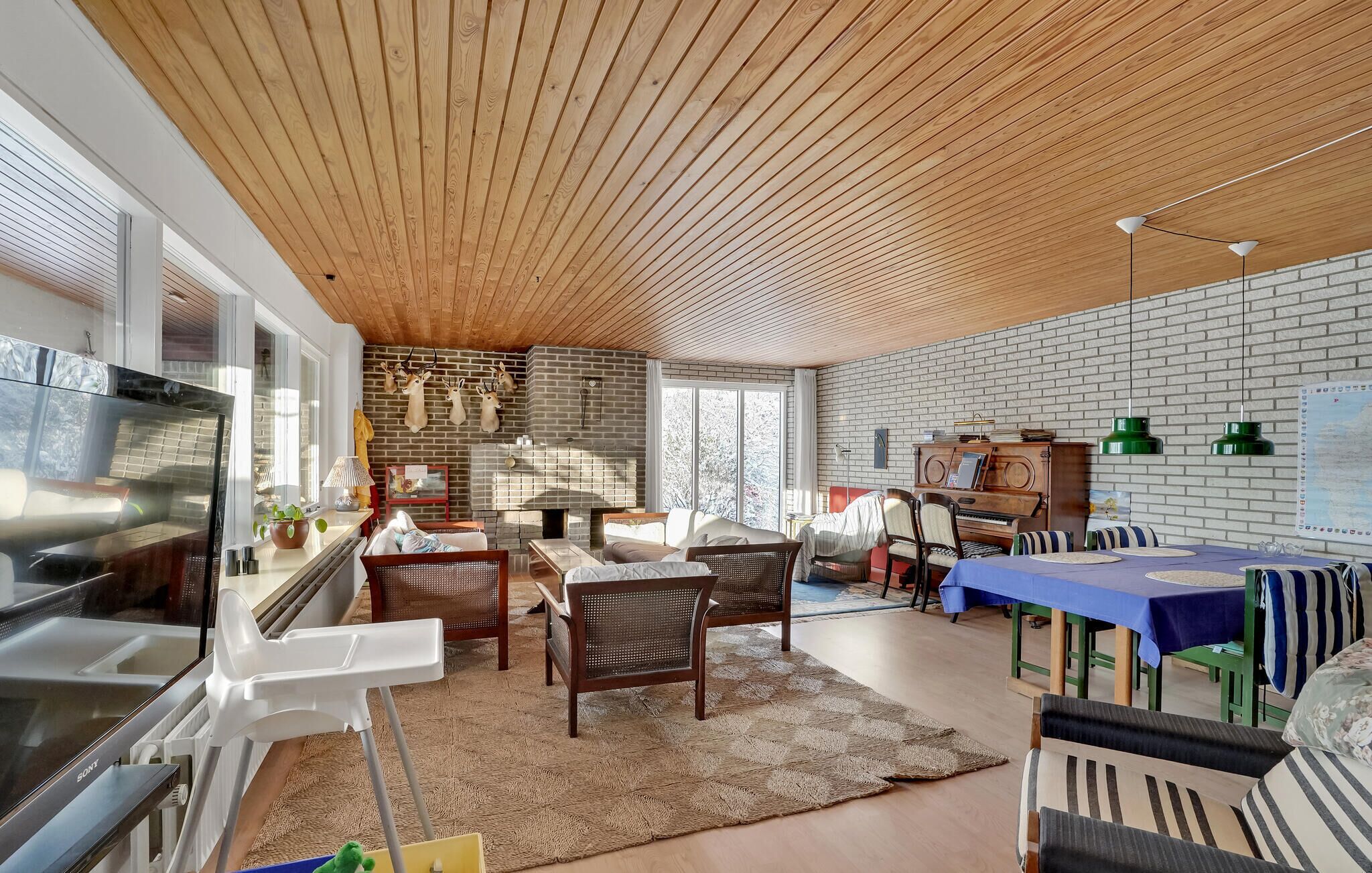 Una sala de estar moderna con techo de madera, pared de ladrillo y chimenea. Hay sofás, sillones y una mesa de comedor.