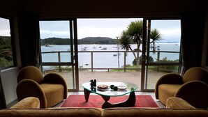 A room with a large window overlooking a body of water with boats. There is a glass coffee table with a red rug, two brown armchairs, and a wooden deck outside. - Greenvale Bed and Breakfast (Oban)