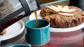 A coffee pot pouring coffee into a blue mug, a plate with cookies and apple slices, and a white plate with a piece of bread. - Greenvale Bed and Breakfast (Oban)