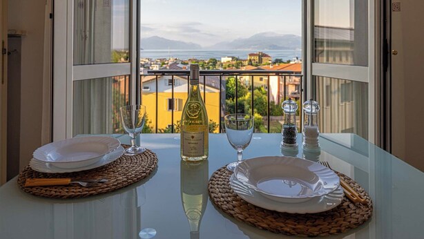 A balcony table set with plates, a wine bottle, and glasses, overlooking a cityscape with buildings and mountains. - Exclusive apartment a stone's throw from the station spectacular lake view Daisy  (Desenzano del Garda)
