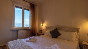 A neatly made bed with white linens and towels, a headboard with decorative carvings, a nightstand with a framed picture, and a window with a view of the sea. - Exclusive apartment a stone's throw from the station spectacular lake view Daisy  (Desenzano del Garda)