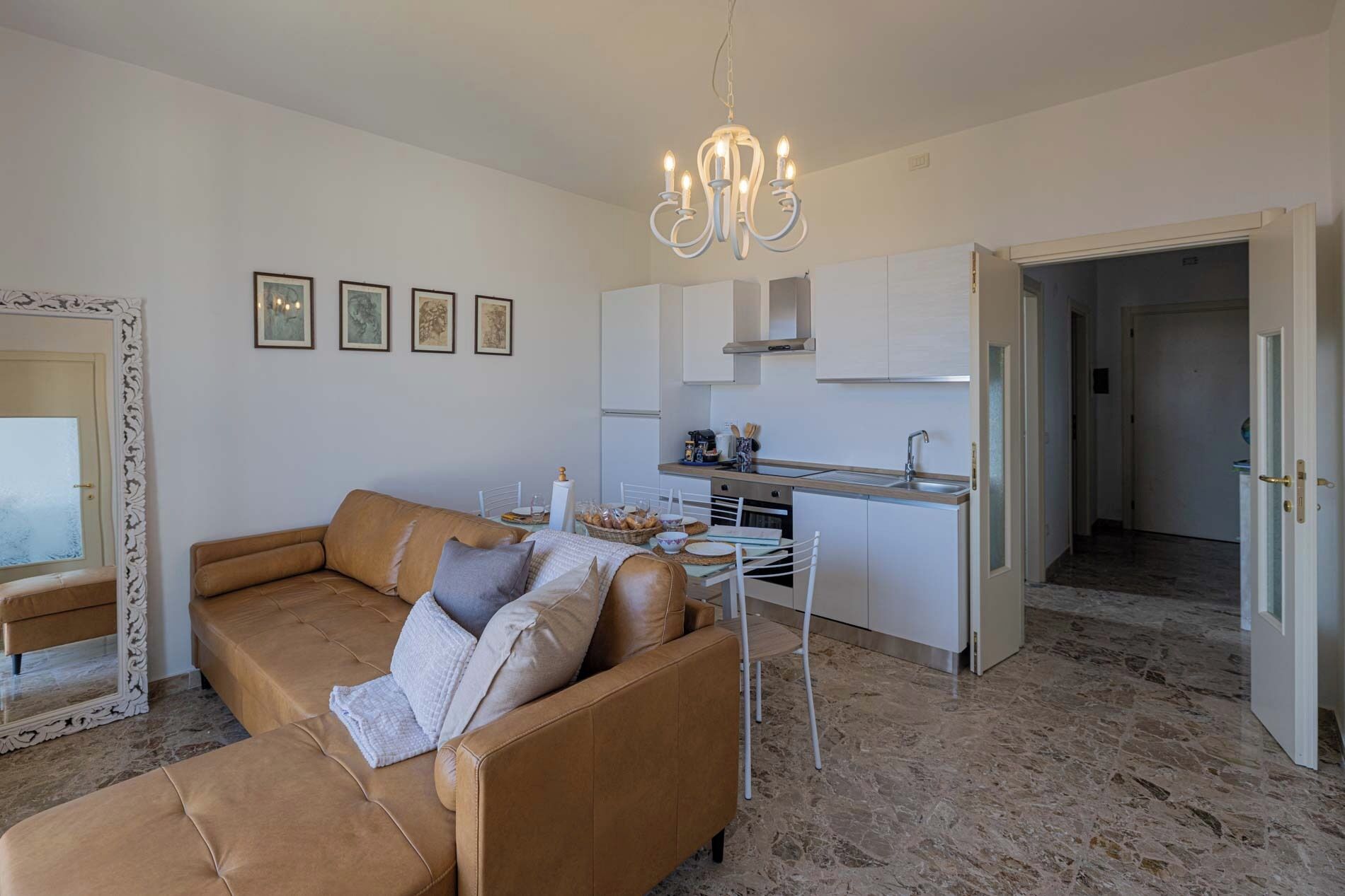 Un soggiorno moderno con un divano in pelle marrone, un tavolo da pranzo con sedie, una zona cucina con mobili bianchi e un lampadario.