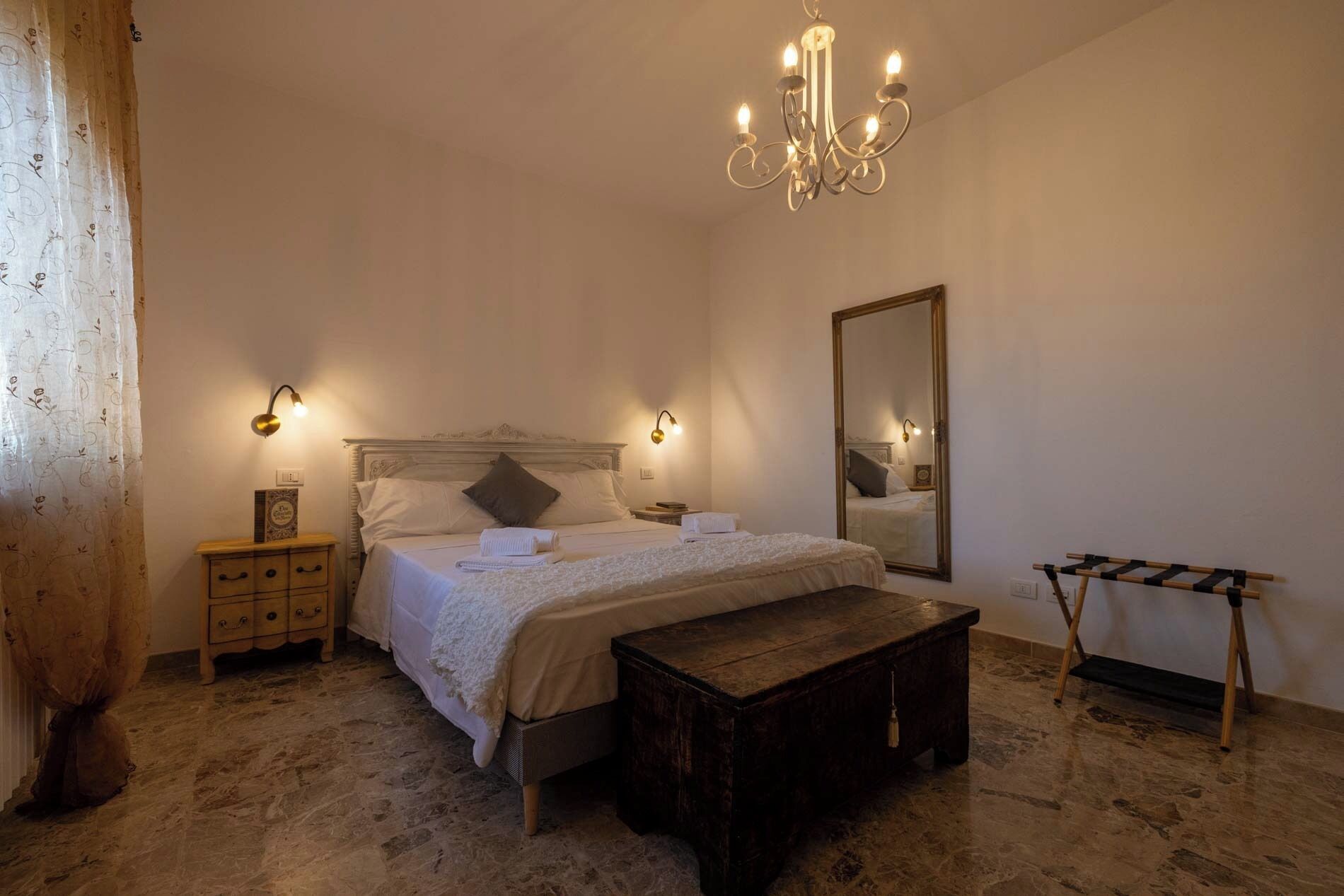 A bedroom with a large bed, bedside tables, a wooden bench, a mirror, and a chandelier.