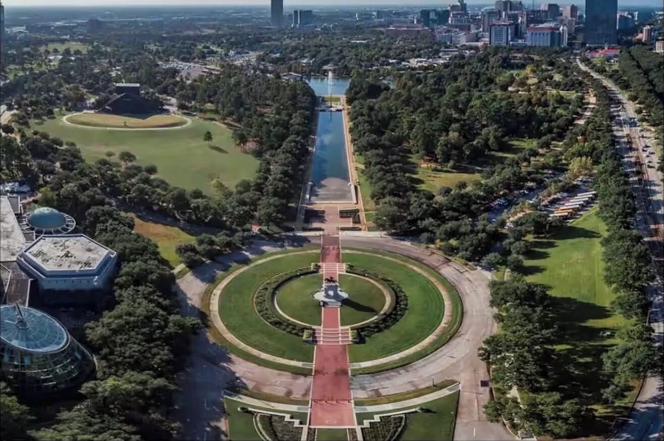 An aerial view of a park with a central pathway, surrounding greenery, and a distant cityscape.