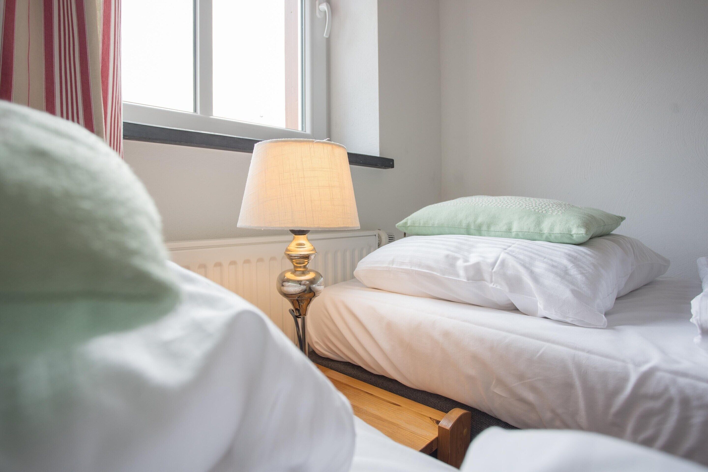 A neatly made bed with white bedding and a green pillow, a bedside lamp, and a window with curtains.
