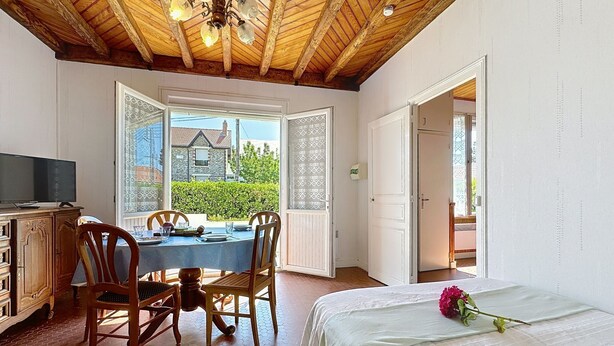 A bedroom with a bed, a dining table with chairs, a TV, and a view of a house through an open door. - Brin de Muguet by Interhome (Tharon Plage)