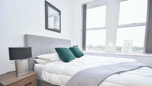A bedroom with a bed, a nightstand, a lamp, a framed picture, and a window with a view of buildings.