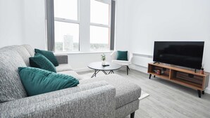A living room with a grey sofa, a white armchair, a round coffee table, a TV on a wooden stand, and a window with curtains.